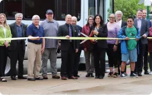 A group of people standing in front of a white bus, smiling as a woman in the center cuts a bright yellow ribbon with large scissors.