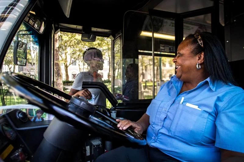 A smiling bus driver in a blue uniform interacts with a passenger through a clear protective barrier.