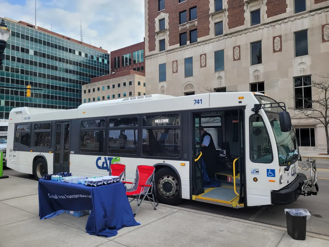 A white CATA bus parked on a city street next to a blue promotional table and red folding chairs.”.  • Ride with our apps: “A woman sitting on a bus, using her smartphone to access a transit app while holding a bus pass.”